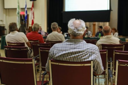 A closeup and back view of an older man sitting indoors during a city hall legislation meeting, blurry attendees are seen in background with copy-space.